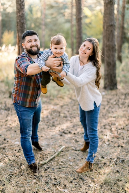 Happy young parents and kid little son, walking together outdoor in autumn forest or park, holding their baby on hands and posing to camera with smile