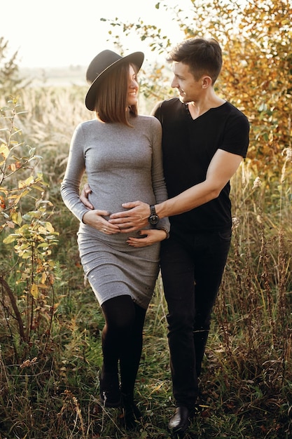 Happy young parents mom and dad hugging baby bump walking and smiling enjoying beautiful moment at sunset Stylish pregnant couple holding hands on belly in sunny light in autumn park