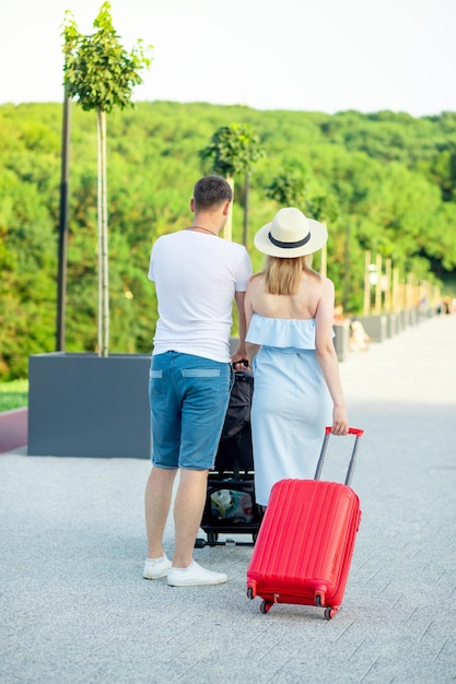 Felici giovani genitori mamma e papà con una valigia rossa e un bambino in un passeggino stanno andando in vacanza in estate la vista dal retro