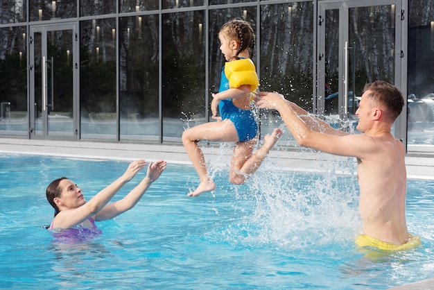 Giovani genitori felici che giocano con la loro piccola figlia carina in piscina davanti alla telecamera mentre trascorrono il fine settimana insieme nel centro termale