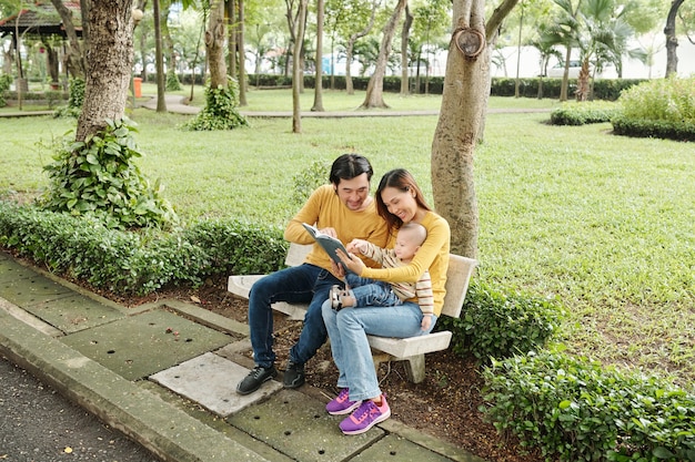 Happy young parents sitting on bench in park and reading interesting book to their little son