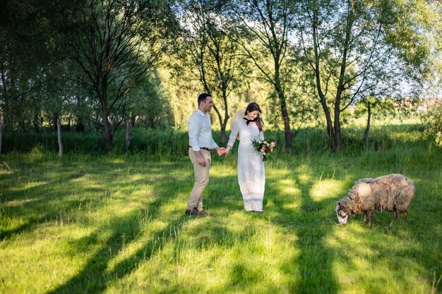 Happy young people in wedding dresses stand in the meadow and look at the lamb