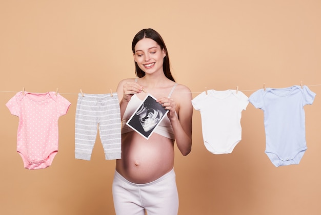 Happy young pregnant girl, with a bared belly, hangs baby clothes on a rope to dry, posing on a beige background. The concept of family, procreation, heir, pregnancy, childbirth, motherhood.