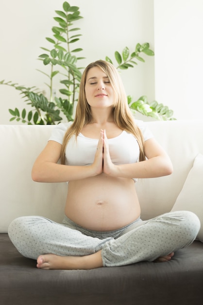 Happy young pregnant woman sitting in lotus position on sofa