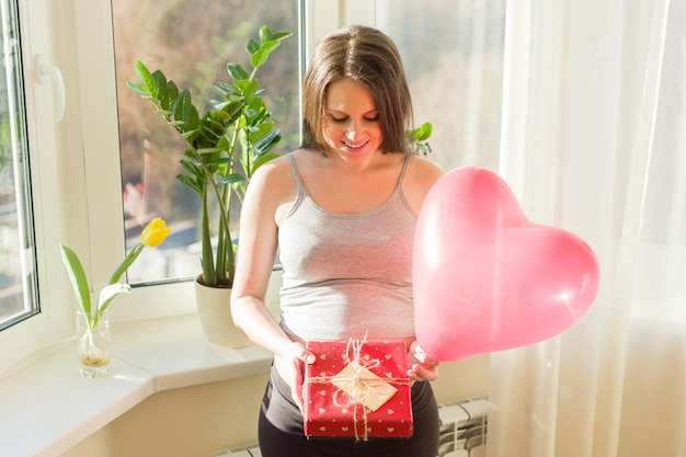 Happy young pregnant woman with gift box and heart balloon