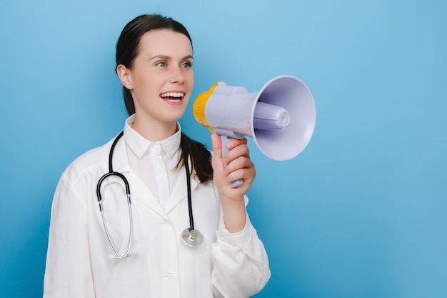 Happy young professional doctor woman in white uniform shouting through megaphone to announce something, posing isolated over pastel blue color background in studio. People emotion lifestyle concept