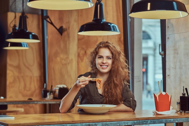 Felice giovane donna rossa che indossa abiti casual che mangiano noodles piccanti in un ristorante asiatico.