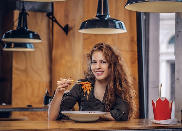 Счастливая молодая рыжая женщина в повседневной одежде ест острую лапшу в азиатском ресторане.