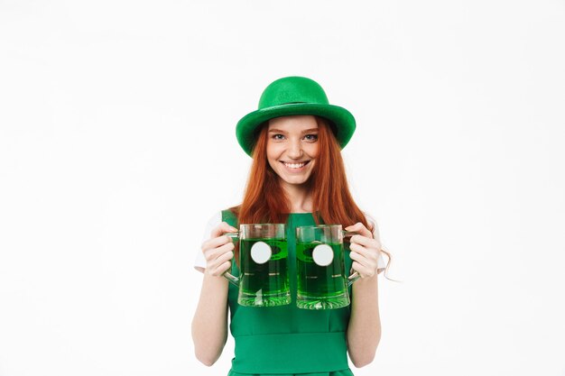 Happy young redheaded girl wearing green hat, celebrating St Patricks's Day isolated over white wall, drinking beer