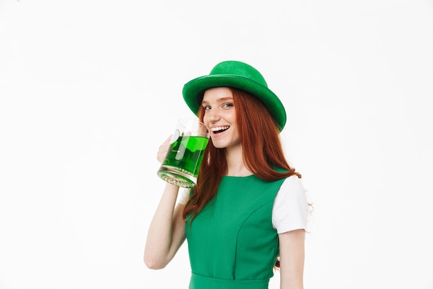 Happy young redheaded girl wearing green hat, celebrating St Patricks's Day isolated over white wall, drinking beer