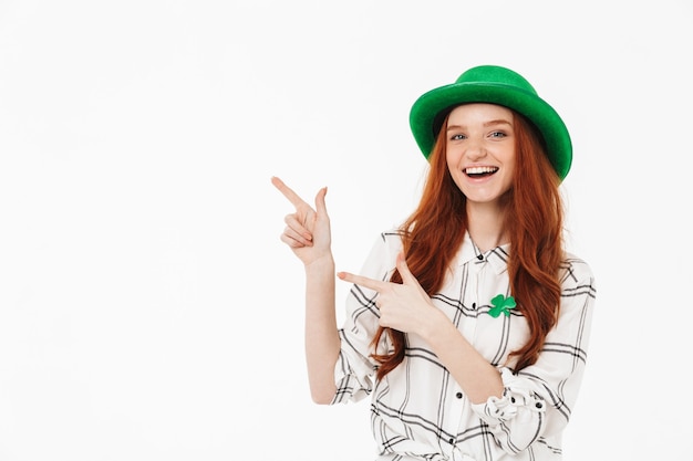 Happy young redheaded girl wearing green hat, celebrating St Patricks's Day isolated over white wall, pointing finger