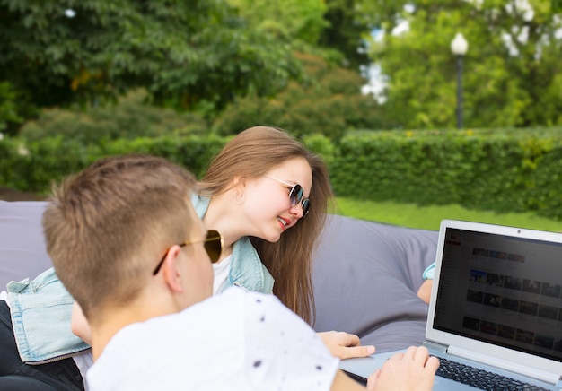 Happy young relaxed couple in sunglasses lying on a big cushion with laptop in park.