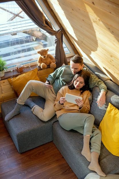 Счастливые молодые расслабленные муж и жена в повседневной одежде лежат на большом диване в своем загородном доме, наслаждаются отдыхом и смотрят онлайн-фильм
