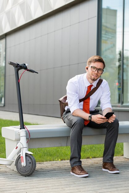 Happy young restful businessman with smartphone sitting on bench against exterior of modern building outdoors