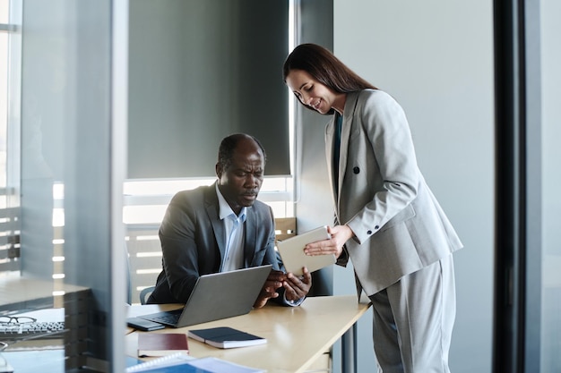 Happy young secretary with tablet bending over workplace of her boss