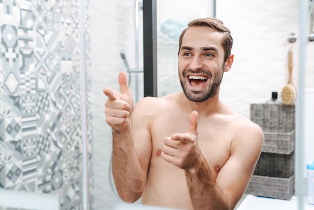 Felice giovane uomo a torso nudo che si guarda allo specchio del bagno Foto Premium