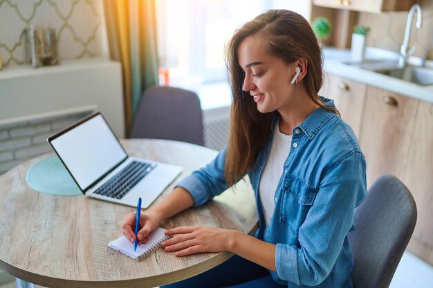 Happy young smart millennial woman takes a note with pen in a notepad and using laptop with open white blank display for online education at workspace. Computer mockup for website advertising