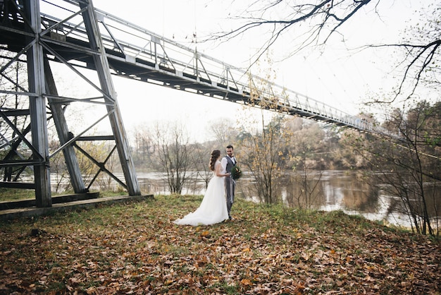 Счастливые молодые улыбающиеся жених и невеста стоят возле висячего моста и реки. Свадебные фото в интересном месте