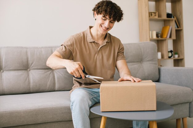 Happy young smiling curly man opening box with ordered goods
gifts presents at home on couch online shopper male customer
opening online shop parcel