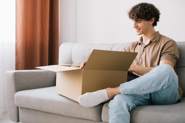 Happy young smiling curly man opening box with ordered goods gifts presents at home on couch Online shopper male customer opening online shop parcel