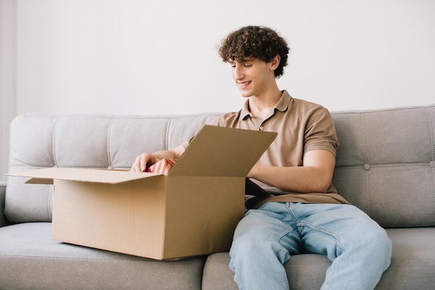Happy young smiling curly man opening box with ordered goods gifts presents at home on couch Online shopper male customer opening online shop parcel