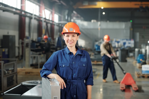 Happy young smiling female engineer in hardhat and blue coveralls looking at you while standing in warehouse of factory