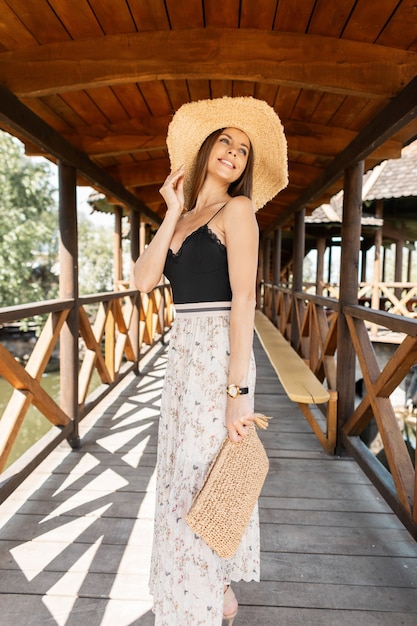 Happy young smiling woman with slim body in wicker straw hat and fashionable handbag in vintage lace dress is walking in nature near the beach