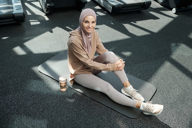 Happy young sportswoman in hijab and activewear looking at you while sitting on mat in gym