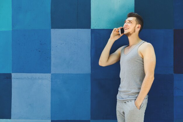 Happy young sporty man talking on phone, having break after training, standing at bright blue graffiti wall, copy space