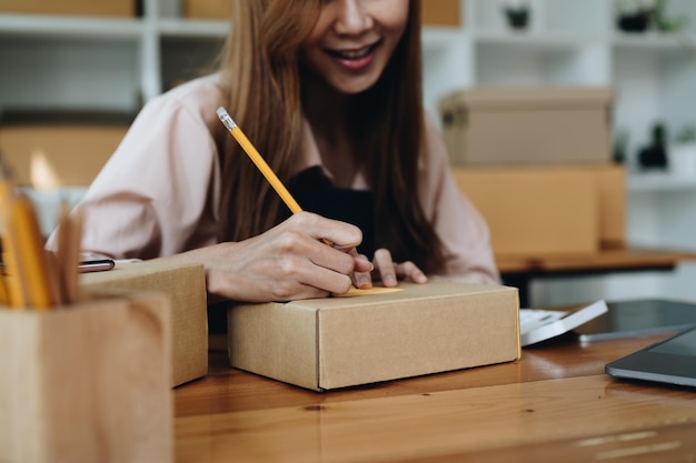 幸せな若いスタートアップの中小企業の所有者は、段ボール箱に住所を書いて梱包しますフリーランスの女性売り手は、顧客に届けるために製品の小包箱を準備しますオンライン販売の配送コンセプト