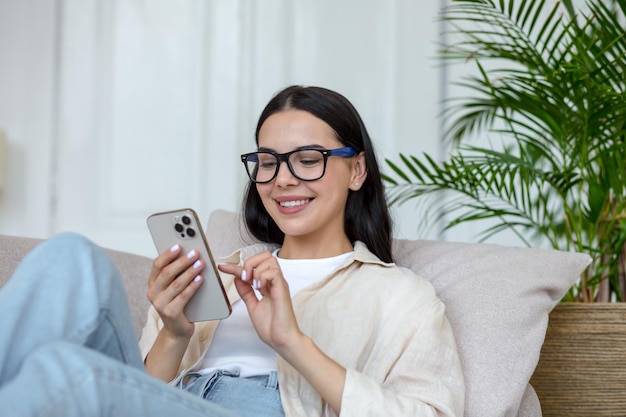 Happy young student girl in glasses sitting on sofa at home holding phone chatting and chatting with