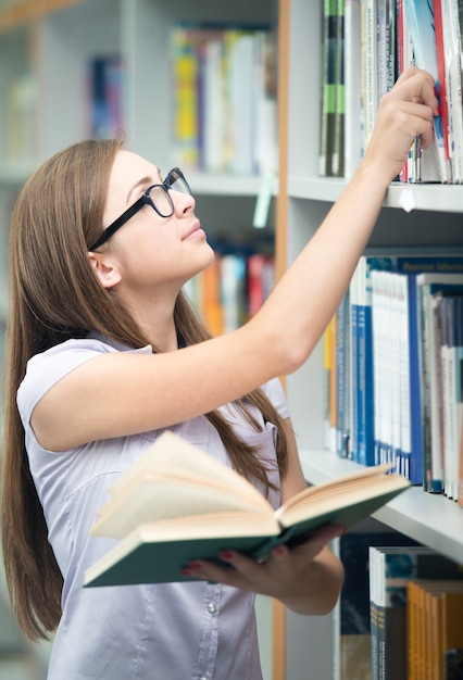 Giovani studenti felici che studiano nella biblioteca universitaria che seleziona un libro dallo scaffale