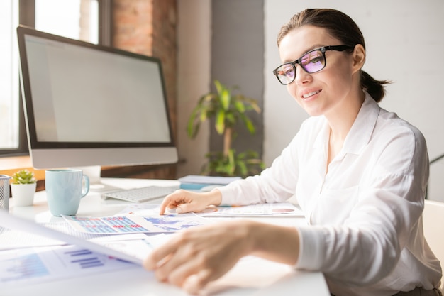 Happy young successful businesswoman sitting by desk with lots of papers in front and reading one of them at work