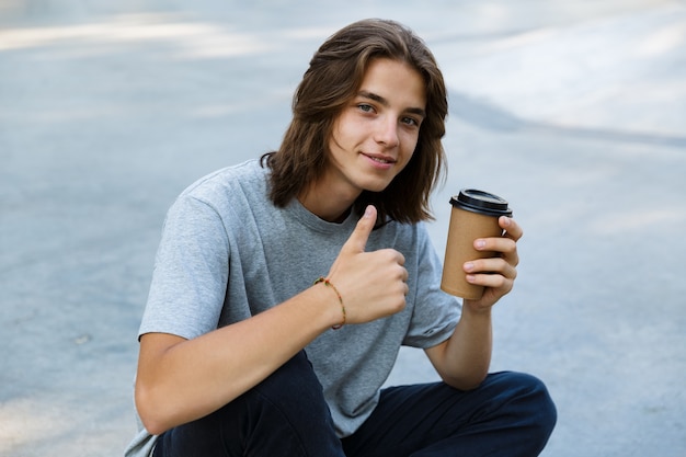 Happy young teenge boy spending time at the skate park, sitting on a skateboard, holding takeaway coffee, giving thumbs up