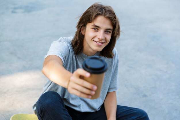 Photo happy young teenge boy spending time at the skate park, sitting on a skateboard, holding takeaway coffee