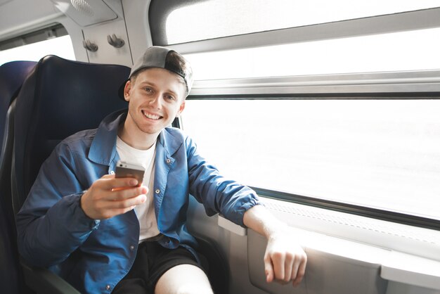 Happy young traveler sitting in a train near the window, using a smartphone