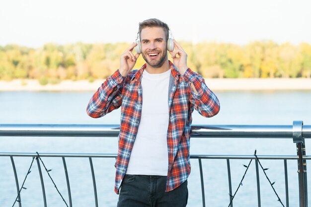 Happy young trendy male fashion model in music headphones and casual shirt outside music