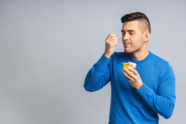 Happy young ukrainian handsome man eating yogurt isolated over\
grey white background healthy lifestyle concept