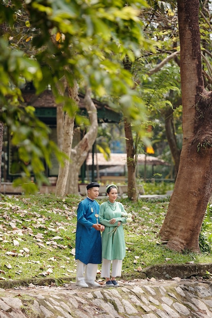 Happy young Vietnamese couple in ao dai dresses looking at pond water in city park spending day together after ceremony