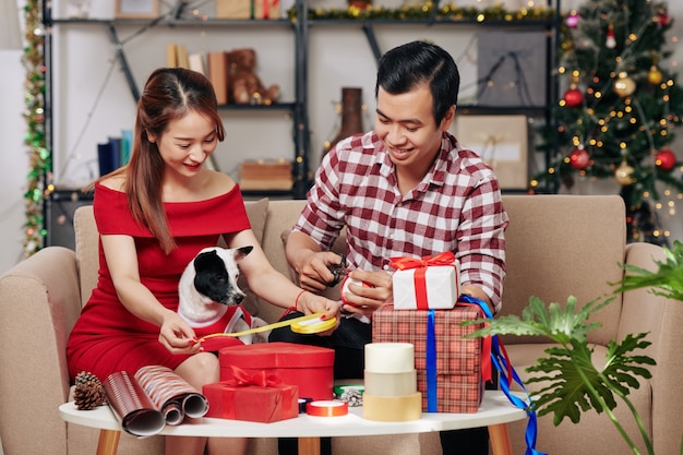 귀여운 작은 개가 화려한 리본을 자르고 크리스마스 선물을 포장하는 행복한 젊은 베트남 커플