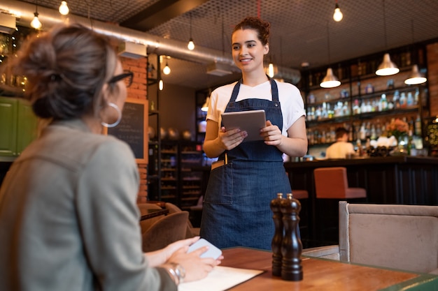 Счастливая молодая официантка с цифровым планшетом стоит у одного из столов перед гостем-женщиной и принимает ее заказ в кафе или ресторане