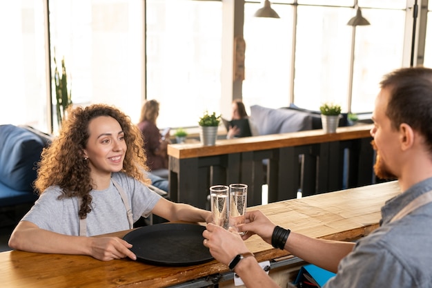 Счастливая молодая официантка с подносом смотрит на своего коллегу у прилавка, ставит два стакана воды для клиентов