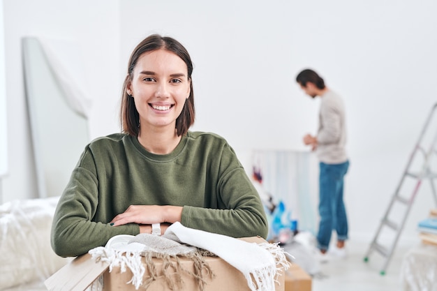 Happy young wife with toothy smile looking at you while unpacking boxes