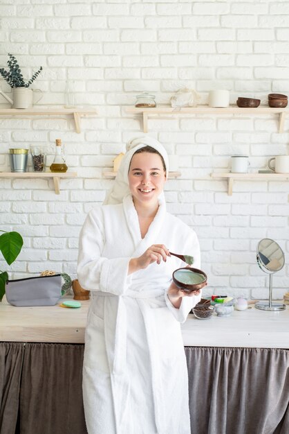 Foto felice giovane donna applicando scrub viso sul viso nella sua cucina di casa