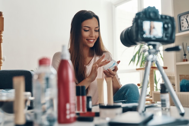 Photo happy young woman applying lip gloss and smiling while making social media video