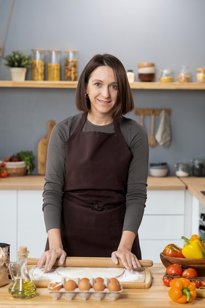 Happy young woman in apron standing by kitchen table