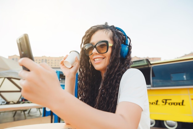 A happy young woman attending a web conference through headphones or a freelancer sitting at an outdoor cafe table a girl with dreadlocks communicates via video link