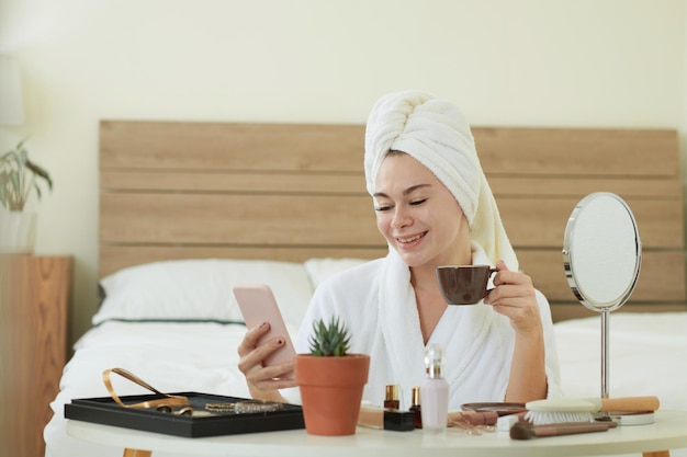 Felice giovane donna in accappatoio che beve caffè e legge messaggi di testo da un amico sullo smartphone