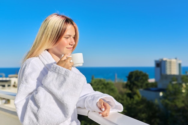 목욕 가운을 입은 행복한 젊은 여성이 커피 한 잔과 바다 리조트의 햇살 가득한 풍경을 즐기고 있습니다.