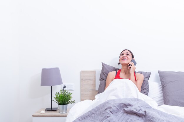 Happy young woman in bed smiling while listening on mobile phone